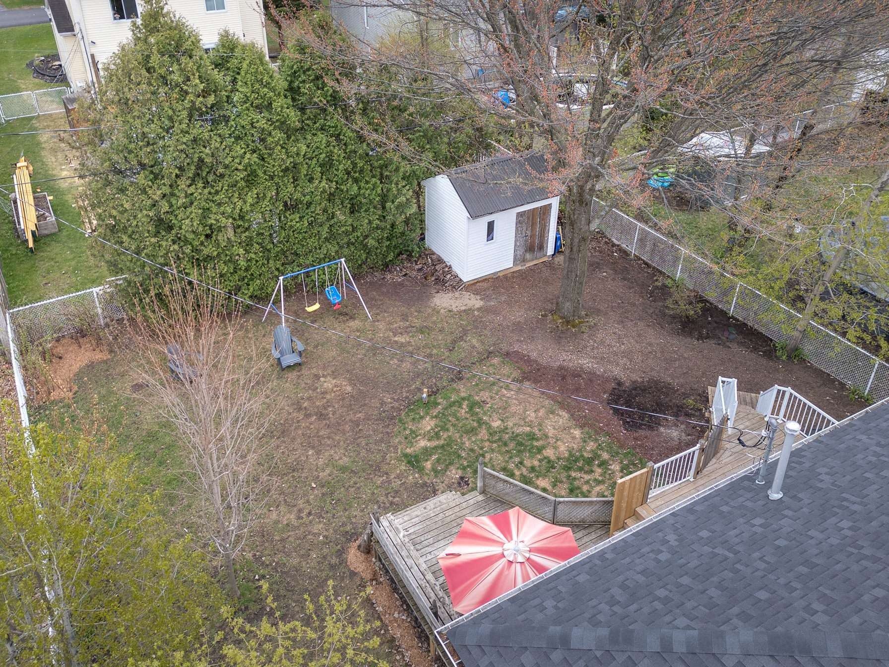 Aerial view of the backyard showing the L-shaped deck, garden shed, mature trees, privacy hedge, swing set, and fenced perimeter