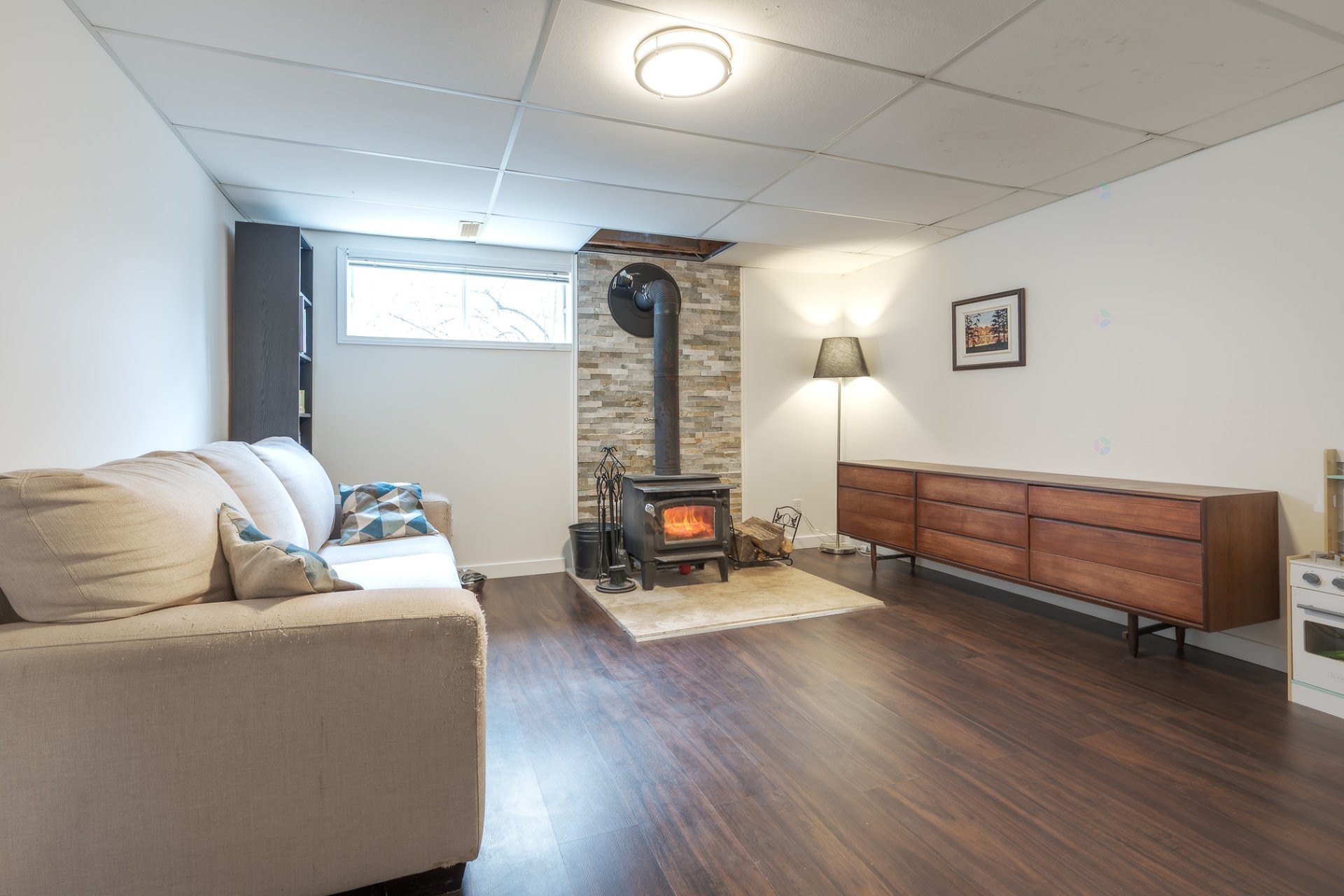 Basement family room from a wider angle showing the wood stove, dark wood floors, midcentury sideboard