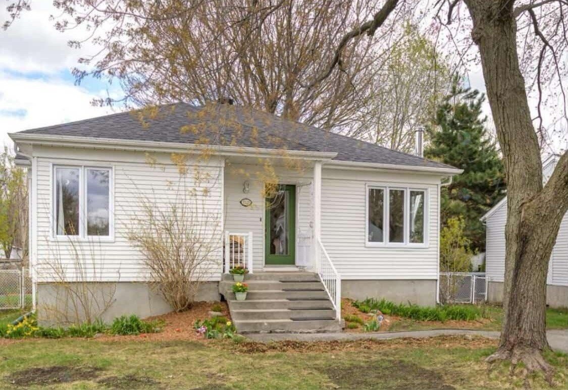 1728 rue des Capucines, front exterior — white siding bungalow with sage green door, mature trees