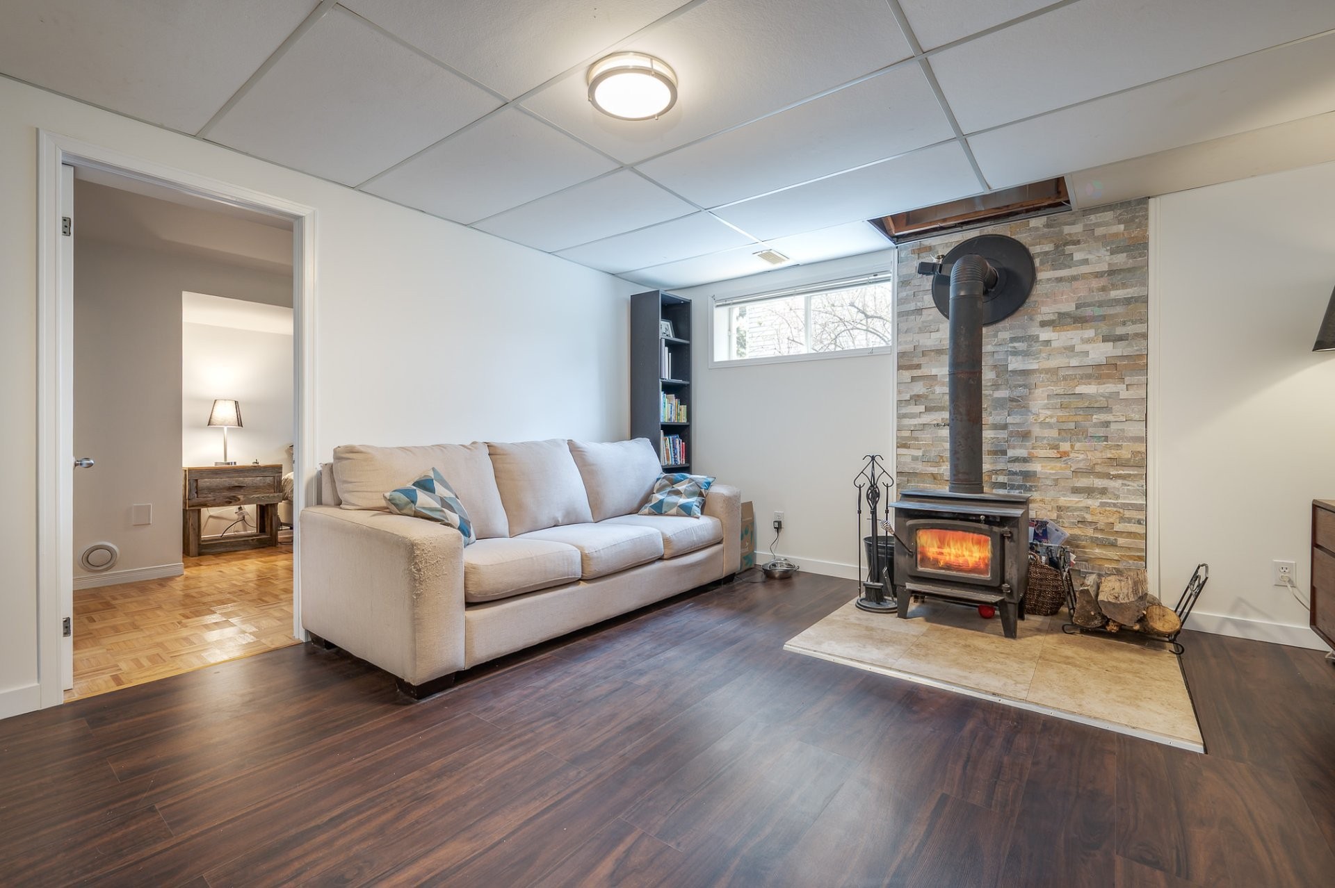 Basement family room with a wood stove lit, stone surround, white couch, view through doorway to the basement bedroom